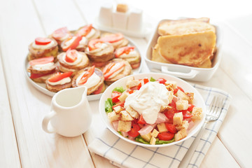 fried zucchini with tomatoes and sour cream sauce; salad with cheese, ham, toast and herbs on a white wooden table