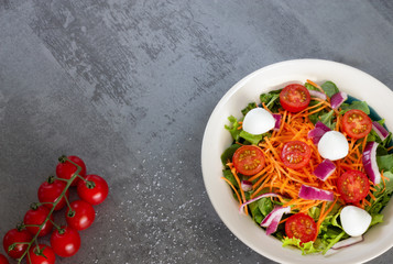 green leaf salad with carrot, tomato, onion and cheese on gray table with salt and tomatoes top view