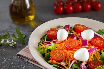 close up green leafy salad with carrot, tomato, onion and cheese on gray table with tomatoes and olive oil in the background