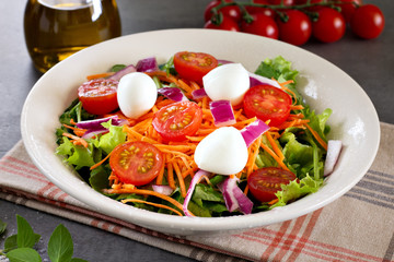 close up green leafy salad with carrot, tomato, onion and cheese on gray table with tomatoes and olive oil in the background