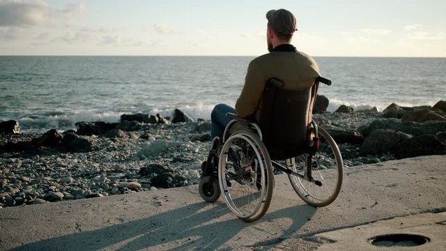 Lonely man is sitting in invalid carriage on sea shore, looking in distance