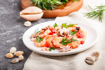 Boiled shrimps or prawns and small octopuses on white ceramic plate on a black concrete background and linen textile. side view, close up.