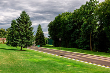 asphalt road with yellow marking of the bike path in the park with green grass and trees lit by sunlight after rain with a cloudy sky.