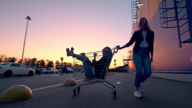 Two millennials girlfriends in street clothes have fun in a supermarket parking lot at sunset. Riding a shopping cart, enjoying freedom.