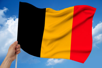 hand holds against the background of the sky with clouds the colored flag of Belgium on the texture of the fabric, silk with waves, close-up