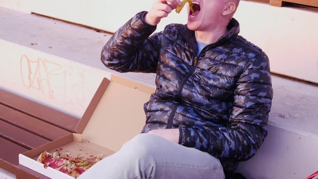 Young bald man sitting in a Park eating pizza out of a box. He eats hot pepper, winces and waves his hands