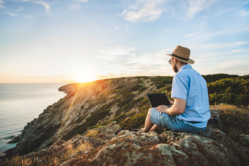 Male travel blogger sitting on a cliff with his laptop