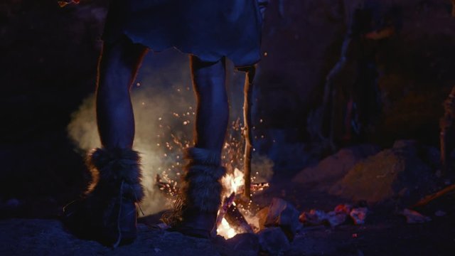 Close-up prehistoric tribe leader legs standing at bonfire warming up near cave dwelling. First human society. Era of ice age.
