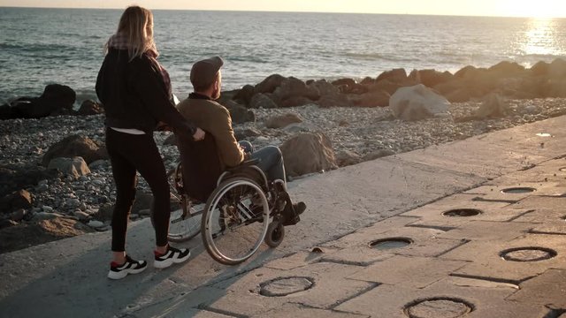 Young nurse is rolling invalid carriage with adult man over sea shore