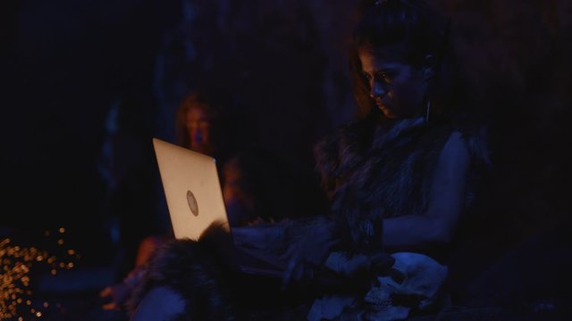 Ancient smart business woman in animal skin using modern laptop computer at bonfire. Caveman tribe leader in background. Homo sapiens.