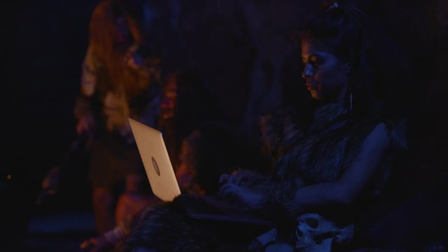 Savage neanderthal woman using modern computer laptop like modern girl annoyed by stupid cavemen playing with fire outside cave at night. Earliest IT technologist.
