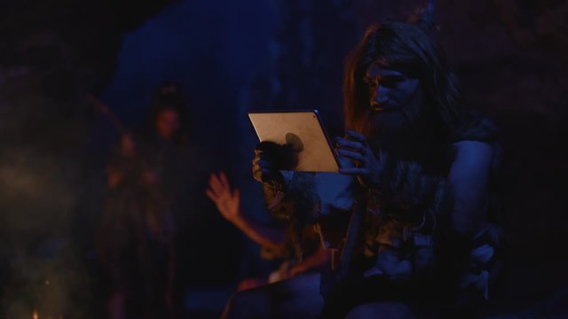 Portrait of baffled savage caveman typing hard on modern tablet future device. Primal tribe at bonfire. Evolution and education.
