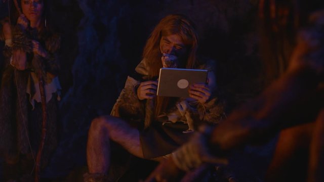 Prehistoric wild tribe leader eating raw meat and using mobile smart tablet outside cave at bonfire on dark night. Evolution of homo sapiens.