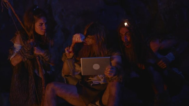 Group of wild nomadic savages using future gadget with surprise and fun warming up by bonfire outside cave. Neanderthals family. Evolution and technology.