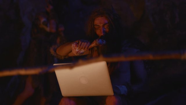 Prehistoric cro-magnon IT technologist using laptop computer at bonfire. Portrait of ancient primitive man discovering technology. Homo sapiens.