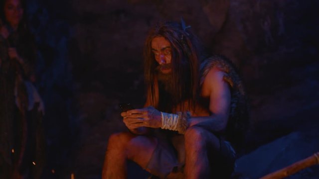 Primitive wildman of paleolithic period outside cave dwelling tapping on new toy future smartphone technology at night. Cro-magnon human tribe at bonfire. Homo sapiens.
