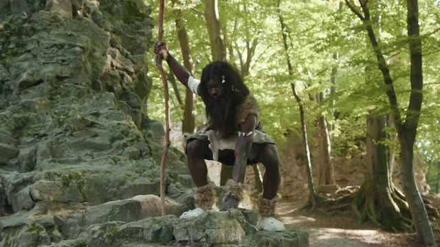 Wild african savage in animal fur costume sitting on rocks watching animals food hunting in the forest near cave. Aboriginal tribe leader. Homo sapiens.