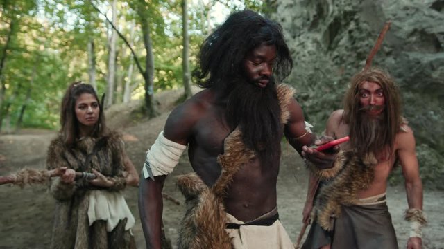 Group of primitive savages hunting animals in forest. African tribe leader using smartphone talking with friend on future gadget. Technology in prehistory.