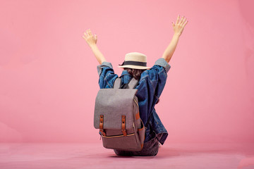 Smiling woman traveler sitting raising arms with backpack in holiday on pink backgrounds,...