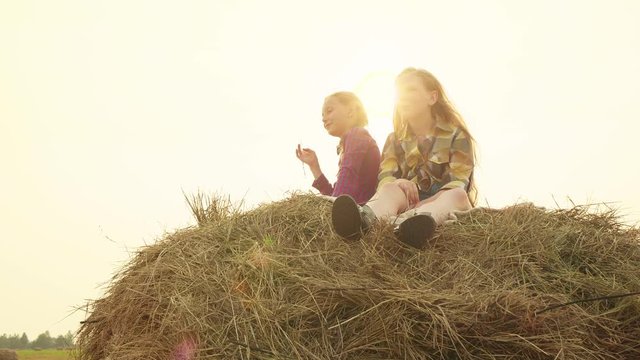 Two teenager girls sitting on hay stack on evening sunset background. Girl teenagers relaxing on haystack at harvesting field. Girl resting on haystack in village while golden sunset