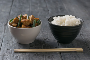 A bowl of boiled rice and a bowl of fried tofu cheese on a wooden table.