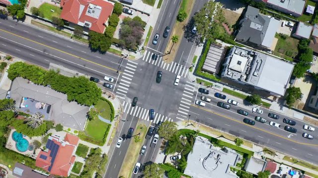 Los Angeles. Aerial. 4K. Drone rises above the intersection of the roads in a suburb. Cars drive in different directions. The roofs of the houses, pools and trees are around.