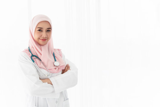 a beautiful young Islamic doctor woman with headphones stood across her arms and smiled confidently in the examination room near the window with curtains and white light from outside. Portrait photo.