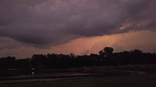 A car with its lights on slowly drives around a lake, as lightning flashes in the background and dusk turns to dark. Trees are silhouetted on the horizon as ominous looking clouds role across the sky.