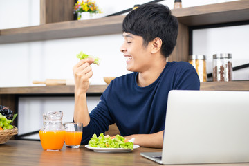 Smiling young Asian businessman working on laptop and having a fresh vegetable salad, mixed fruit and orange juice for lunch break. Healthy food, eating right and people lifestyle concept.