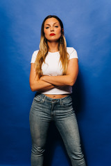 Attractive young woman standing in a serious way while looking at the camera with her hands between loops wearing white shirt and blue pants with blue background.