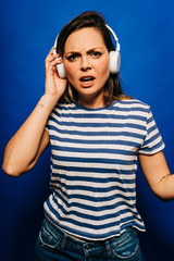 Beautiful young blonde listening to music with expression of doubt wearing striped shirt and blue pants with blue background.