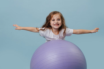 Little girl is trying to climb on a yoga ball without hands over blue background