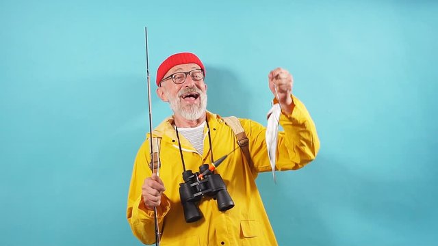 Happy old bearded fisherman with binocular wearing red casual hat and yellow anorak holding in hand fish which he catched on sea last night. good day for fishing, positive feeling and emotion