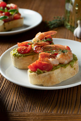 bruschetta with bell shrimp, avocado, pesto sauce and cherry tomato on a white plate on a wooden background. Italian restaurant. Close up food photo
