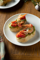 bruschetta with bell shrimp, avocado, pesto sauce and cherry tomato on a white plate on a wooden background. Italian restaurant. Close up food photo