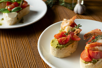 bruschetta with bell shrimp, avocado, pesto sauce and cherry tomato on a white plate on a wooden background. Italian restaurant. Close up food photo