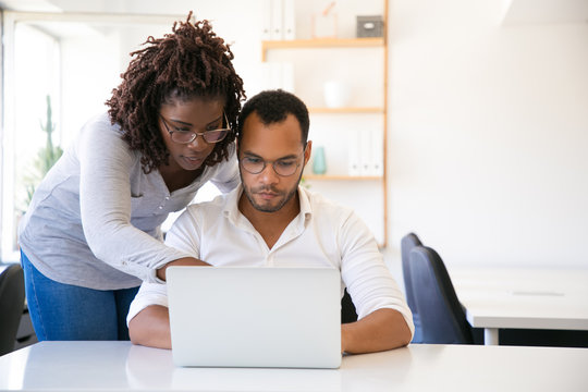 Professional helping new employee with report. Business man and woman in casual sitting and standing at workplace, using laptop, pointing at screen. Mentorship concept