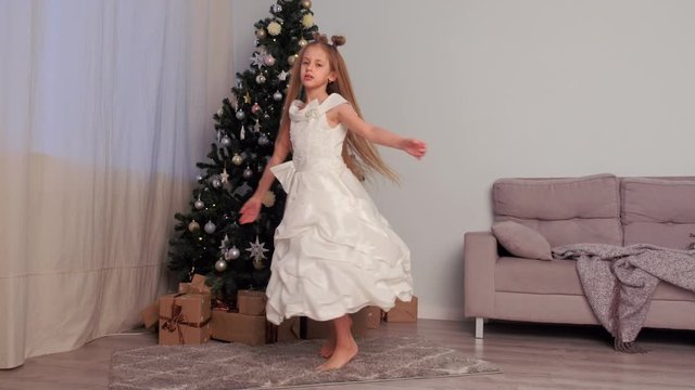 Barfooted beautiful girl with long hairs teen dancing in white dress near Christmas tree at home. Xmas winter holidays. Traditional New Year interior living room. Presents under artificial pine tree.