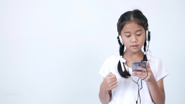 The girl looks cute and wears a white shirt and a braid tied with a white ribbon using the headphones, listening to music from social media on smartphone, singing softly and rocking along the song.
