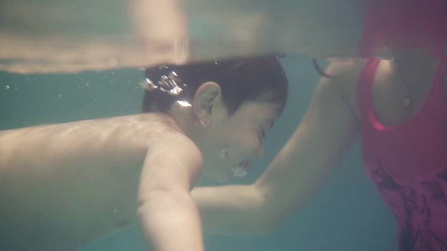 A concentrated brunette woman teaches her child to dive into the water, swimming in the sports pool. Mother and son swim in the pool, the child dives into the water while mother isholding him and