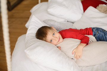 handsome light-eyed boy in red jumper lies among white pillows and blankets