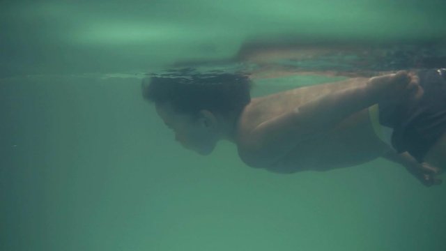 A small swimmer diving into the water and rows with legs under the water, heading to his mother. The boy sits on the edge of the pool, his mother waiting for him in the water. Indoors. Underwater