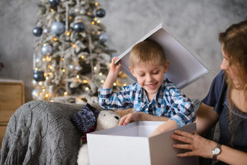 Fototapeta na wymiar boy was delighted and surprised at gift he saw in big gray box, in his mother’s hands, sitting on sofa against background of Christmas tree glowing with them bokeh