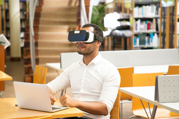 Male student using VR headset during work in library. Latin man wearing virtual reality glasses, sitting at desk with laptop. Education concept
