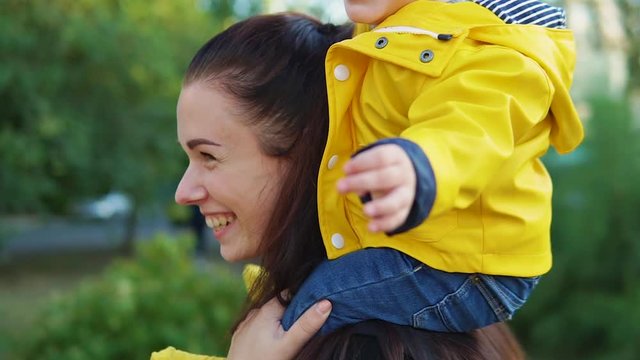Mom plays with her child in the park at sunset. The boy sits on his shoulders in a yellow jacket and smiles happily. Spin together.