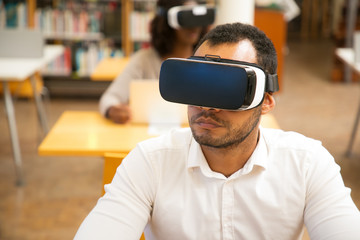 Adult male student using VR glasses while working in library. Latin man wearing virtual reality headset, sitting at desk, enjoying experience. VR gadgets concept