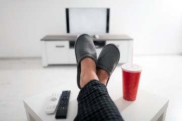 Man drinking soda juice and looking at TV with legs on the table in living room.