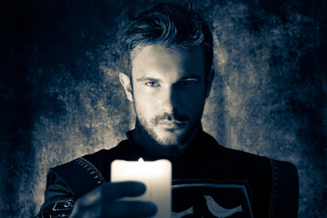 Atmospheric portrait of handsome knight with beard, looking at and holding candle up to camera while casting shadows © Tony Marturano