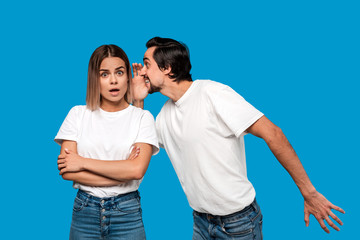 Bearded man with mustaches in white t-shirt tells secret to a young blond girl in white t-shirts and blue jeans standing isolated over blue background. Woman looks surprised. Secret telling.