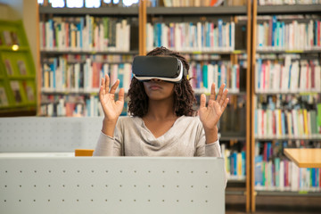 African American student working with VR simulator in library. Young black woman in virtual reality glasses sitting at desk and touching air. VR goggles for studying concept